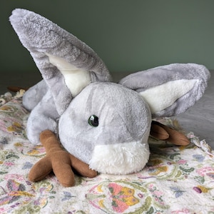 May include: A gray plush bunny with brown felt feet and a white face. The bunny has one green eye and is lying on a floral patterned blanket.