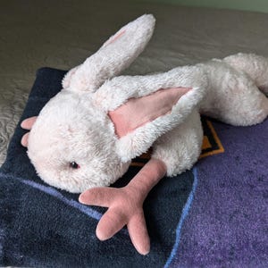 May include: A plush pink bunny with long ears and pink feet. The bunny is lying on a purple and black blanket.