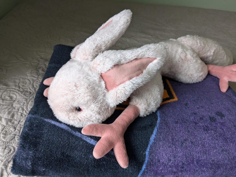 May include: A plush pink bunny with long ears and pink feet. The bunny is lying on a purple and black blanket.