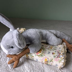 May include: A gray plush bunny toy with long ears and a brown antler-shaped tail. The bunny is lying on a floral patterned pillow with a fringe border.