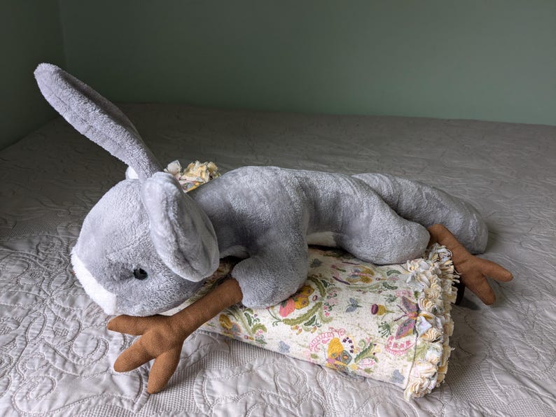 May include: A gray plush bunny toy with long ears and a brown antler-shaped tail. The bunny is lying on a floral patterned pillow with a fringe border.