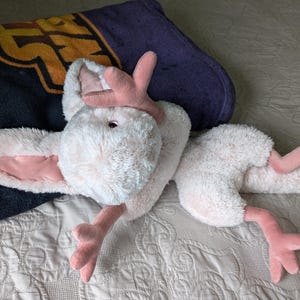 May include: A plush white and pink toy with long ears and a long tail. The toy is lying on a white and gray patterned blanket.