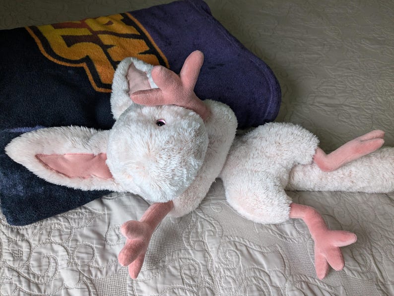 May include: A plush white and pink toy with long ears and a long tail. The toy is lying on a white and gray patterned blanket.