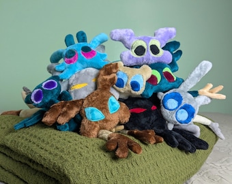 May include: A collection of plush toys in various colors including blue, brown, purple, black, and grey. Each soft toy features large, distinct eyes and multiple limbs or appendages, arranged in a cluster with varied textures and shapes.