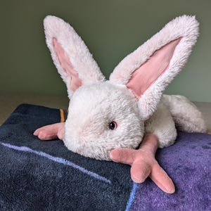 May include: A plush pink and white rabbit with long ears and a fluffy texture. The rabbit is lying on a dark blue and purple blanket.