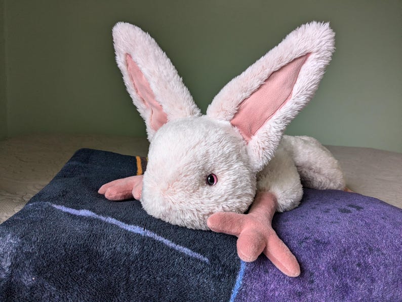 May include: A plush pink and white rabbit with long ears and a fluffy texture. The rabbit is lying on a dark blue and purple blanket.