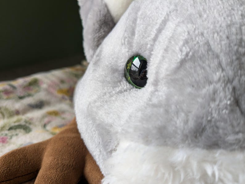 May include: Close-up of a plush toy's eye. The eye is green with a black pupil and a reflective surface.
