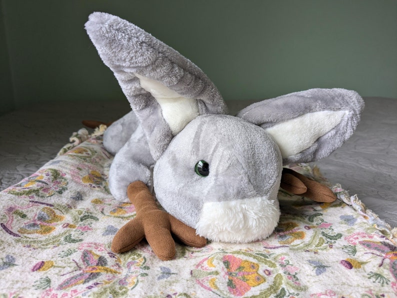 May include: A gray plush bunny with brown felt feet and a white face. The bunny has one green eye and is lying on a floral patterned blanket.