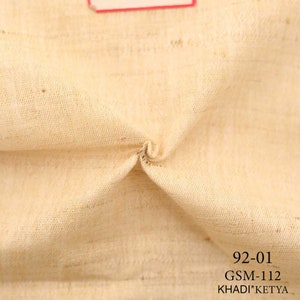 May include: Close-up of a beige fabric with a textured weave. The fabric is folded in the center. The text "92-01 GSM-112 KHADI*KETYA" is printed at the bottom of the image.