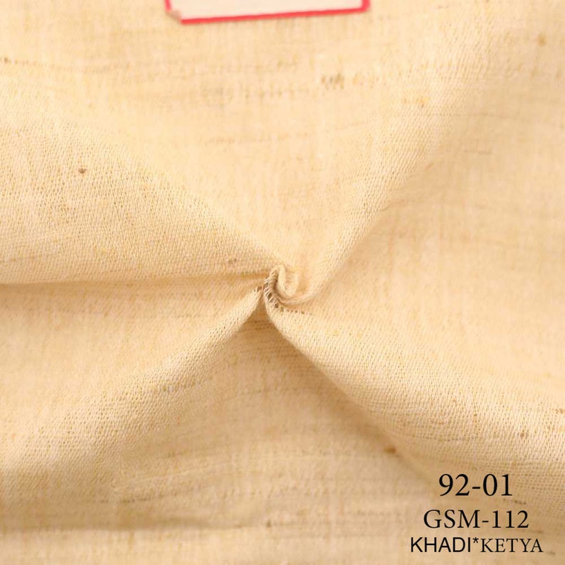 May include: Close-up of a beige fabric with a textured weave. The fabric is folded in the center. The text "92-01 GSM-112 KHADI*KETYA" is printed at the bottom of the image.
