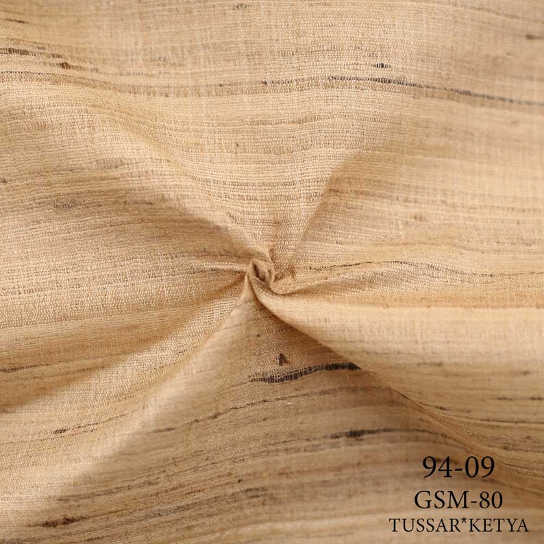 May include: Close-up of a beige fabric with a subtle woven texture. The fabric is folded and has a slight sheen. The text "94-09 GSM-80 TUSSAR*KETYA" is printed in black at the bottom of the image.