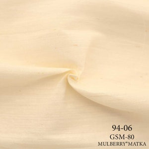 May include: Close-up of a cream-colored fabric with a slightly textured surface. The fabric is folded in the center and has a subtle sheen. The text "94-06 GSM-80 MULBERRY*MATKA" is printed in black at the bottom of the image.