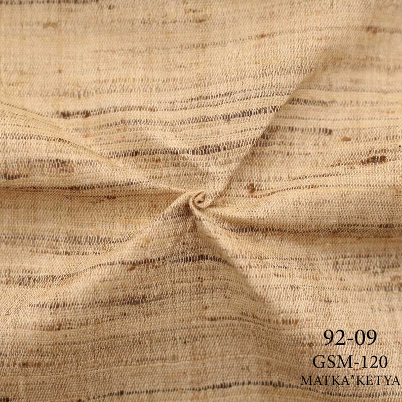 May include: Close-up of a beige fabric with a subtle striped pattern. The fabric is folded and has a rough texture. The text "92-09 GSM-120 MATKA KETYA" is visible in the bottom right corner.