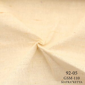 May include: Close-up of a cream-colored fabric with a subtle texture. The fabric has a slightly rough, natural feel. The text "92-05 GSM-110 MATKA*KETYA" is printed in black at the bottom right corner.