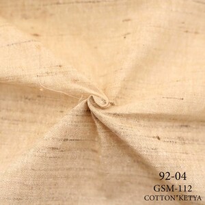 May include: Close-up of a beige fabric with a textured weave. The fabric is folded and has a natural, rustic look. The text "92-04 GSM-112 COTTON*KETYA" is printed in black at the bottom of the image.