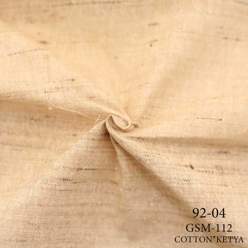 May include: Close-up of a beige fabric with a textured weave. The fabric is folded and has a natural, rustic look. The text "92-04 GSM-112 COTTON*KETYA" is printed in black at the bottom of the image.