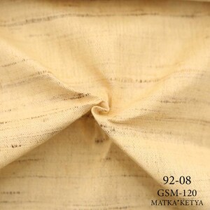 May include: Close-up of a beige fabric with a subtle striped pattern. The fabric is folded and has a soft, textured feel. The text "92-08 GSM-120 MATKA*KETYA" is printed on the fabric.
