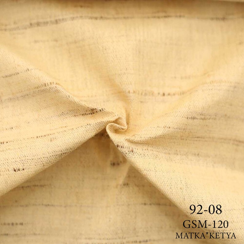 May include: Close-up of a beige fabric with a subtle striped pattern. The fabric is folded and has a soft, textured feel. The text "92-08 GSM-120 MATKA*KETYA" is printed on the fabric.