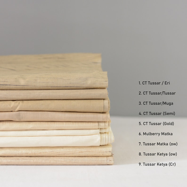 May include: A stack of nine different types of fabric swatches in various shades of beige and cream. The fabric swatches are labeled with their names, including "CT Tussar / Eri", "CT Tussar/Tussar", "CT Tussar/Muga", "CT Tussar (Semi)", "CT Tussar (Gold)", "Mulberry Matka", "Tussar Matka (ow)", "Tussar Ketya (ow)", and "Tussar Ketya (Cr)".