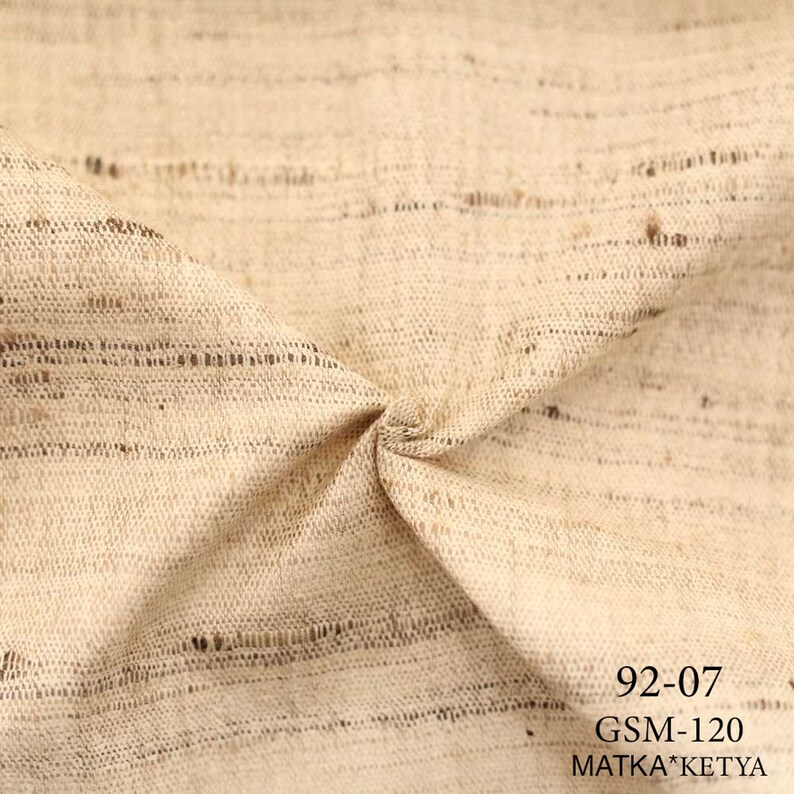 May include: Close-up of a beige fabric with a subtle striped pattern. The fabric is woven with a textured, slightly rough surface. The text "92-07 GSM-120 MATKA*KETYA" is printed in black at the bottom right corner.