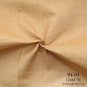 May include: Close-up of a beige fabric with a textured weave. The fabric is folded and twisted, showing its drape and texture. The text "94-03 GSM-70 TUSSA*MUGA" is printed in black at the bottom of the image.