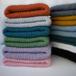 Puede incluir: Una pila de tejido de punto doblado en varios colores, incluyendo naranja, gris, azul verdoso, verde, lavanda y rosa. El tejido está doblado por la mitad y apilado uno encima del otro.