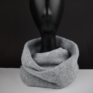 Puede incluir: Una bufanda de punto gris en forma de infinito sobre una cabeza de maniquí negra. La bufanda es suave y acogedora, perfecta para mantenerse caliente en invierno.