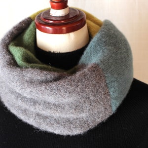 Puede incluir: Una bufanda infinita de punto gris, verde y azul. La bufanda está doblada y se muestra en un maniquí.
