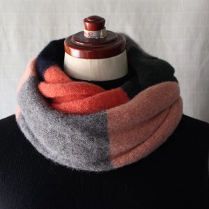 May include: A multi-coloured infinity scarf on a mannequin. The scarf has blocks of coral, grey, navy, and dark green. The scarf is made of a soft, textured material, ideal for colder weather.