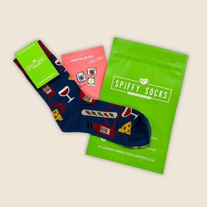 May include: A pair of navy blue socks with wine and cheese graphics, accompanied by a green package and a pink card. The green package has the text "SPIFFY SOCKS". The pink card says "INSTAGRAM CHALLENGE".
