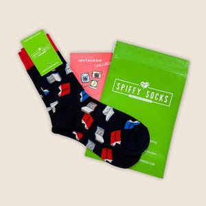 May include: A pair of navy blue socks with a pattern of open books in red, white, and blue. The socks have red cuffs and are packaged with a green bag and a pink card. The green bag has the text "SPIFFY SOCKS".