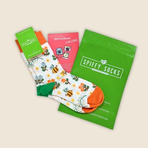May include: A pair of white socks with a bee and floral pattern, orange heels and toes, and green accents. The socks are packaged with a green bag and two cards. The green bag has the text "SPIFFY SOCKS". One card says "SPIFFY SOCKS". The other card says "INSTAGRAM CHALLENGE".