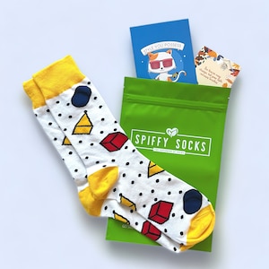 May include: White socks with yellow cuffs and geometric shapes, including triangles, cubes, and circles. The socks are next to a green bag with the text "SPIFFY SOCKS" and a blue card with a cat wearing sunglasses.