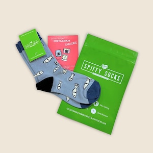 May include: A pair of light blue socks with a fish pattern, a green bag with "SPIFFY SOCKS" printed on it, and a small green card. The socks have navy blue heels and toes. A red card with "INSTAGRAM CHALLENGE" is also visible.