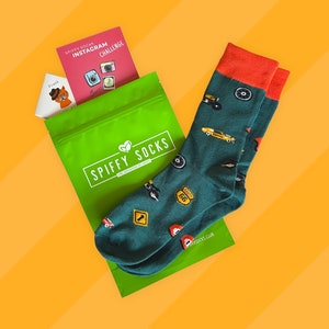 May include: A pair of green and blue socks with a red cuff. The socks feature a variety of graphics, including a Route 66 sign, a monster truck, and a car. The socks are packaged in a green bag with the words "Spiffy Socks" printed on it.