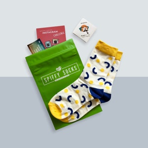 May include: A pair of cream-colored socks with yellow and navy blue geometric patterns, next to a green bag with the text "Spiffy Socks". Also included are small cards with text and illustrations.