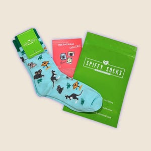 May include: Light blue socks featuring kangaroos, koalas, and road signs, alongside a green package and a pink card. The green package displays "Spiffy Socks". The socks are designed for comfort and style, perfect for everyday wear.