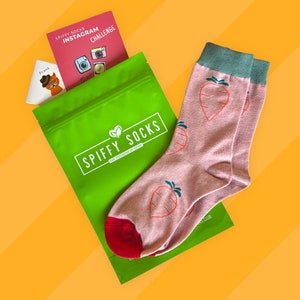 May include: A green pouch with the text "Spiffy Socks" and a pair of pink socks with a green and red carrot design. The socks are folded and the pouch is open.