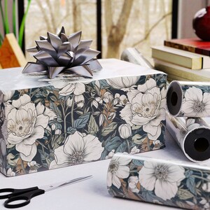 May include: A wrapped gift box with a silver bow, featuring floral patterned wrapping paper in white, navy, and tan. Rolls of matching wrapping paper and a pair of black scissors are also visible.