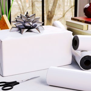 May include: A wrapped white gift box with a silver bow on top. Rolls of white wrapping paper are next to the box, along with a pair of black-handled scissors. The scene is set on a white surface.