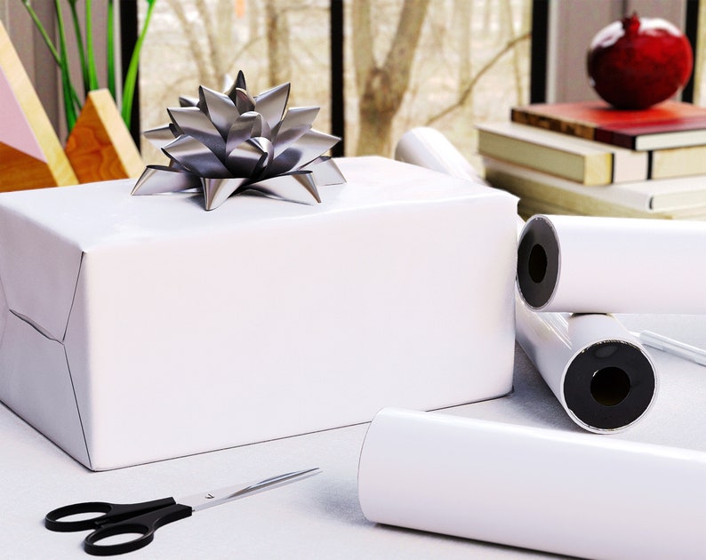 May include: A wrapped white gift box with a silver bow on top. Rolls of white wrapping paper are next to the box, along with a pair of black-handled scissors. The scene is set on a white surface.