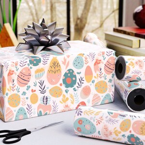 May include: A wrapped gift box with a silver bow, rolls of matching wrapping paper, and scissors. The wrapping paper features a pastel Easter egg and floral pattern. The scene is set on a white surface near a window.