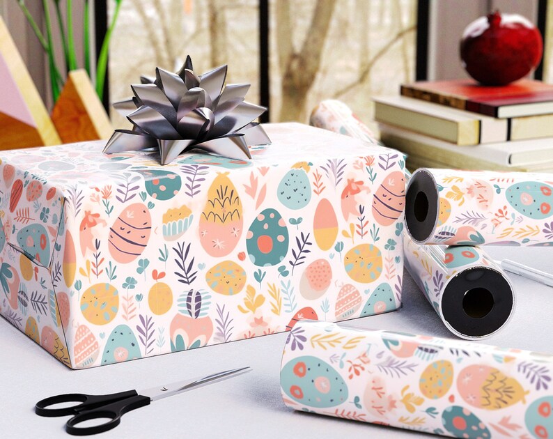 May include: A wrapped gift box with a silver bow, rolls of matching wrapping paper, and scissors. The wrapping paper features a pastel Easter egg and floral pattern. The scene is set on a white surface near a window.