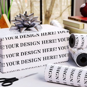 Puede incluir: Un regalo envuelto con un lazo plateado, rollos de papel de regalo a juego y unas tijeras. El papel de regalo es blanco con el texto "YOUR DESIGN HERE!" impreso en negro. La escena está ambientada sobre una superficie blanca.