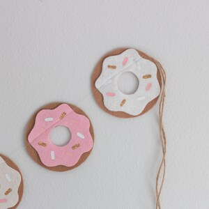 K&ouml;nnte beinhalten: Drei Filz-Donut-Anh&auml;nger mit rosa und wei&szlig;em Zuckerguss und goldenen Streuseln. Die Donuts sind mit einer Schnur aneinandergereiht.
