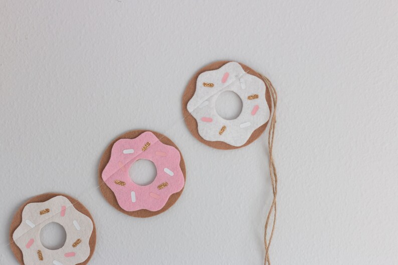 K&ouml;nnte beinhalten: Drei Filz-Donut-Anh&auml;nger mit rosa und wei&szlig;em Zuckerguss und goldenen Streuseln. Die Donuts sind mit einer Schnur aneinandergereiht.
