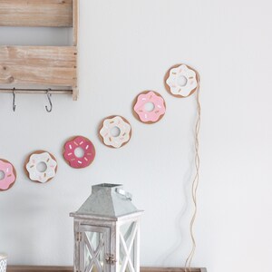 K&ouml;nnte beinhalten: Eine wei&szlig;e Holzlaterne mit einem Metalldach und einer Glasscheibe. Die Laterne steht auf einem Holztisch vor einer wei&szlig;en Wand, an der eine Girlande aus rosa und wei&szlig;en Filzdonuts h&auml;ngt.