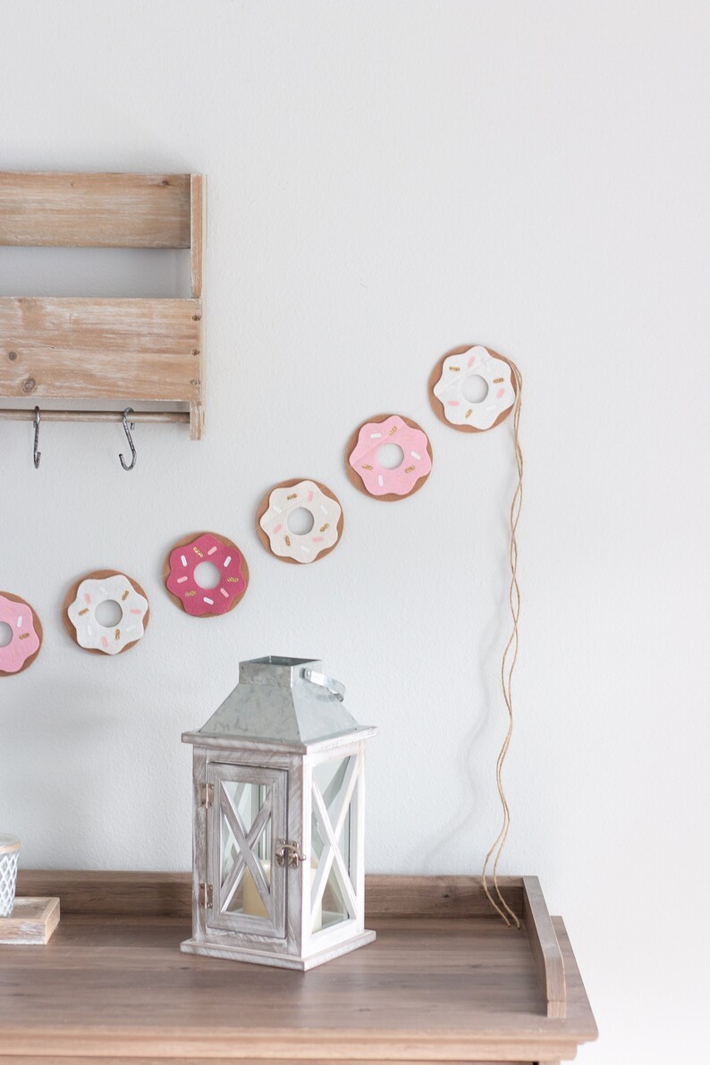 K&ouml;nnte beinhalten: Eine wei&szlig;e Holzlaterne mit einem Metalldach und einer Glasscheibe. Die Laterne steht auf einem Holztisch vor einer wei&szlig;en Wand, an der eine Girlande aus rosa und wei&szlig;en Filzdonuts h&auml;ngt.
