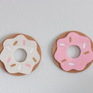 K&ouml;nnte beinhalten: Eine Filz-Donut-Girlande mit rosafarbenen, wei&szlig;en und braunen Donuts. Die Donuts haben goldene Streusel und wei&szlig;es Frosting.