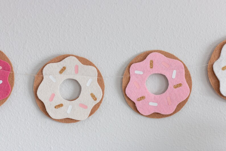 K&ouml;nnte beinhalten: Eine Filz-Donut-Girlande mit rosafarbenen, wei&szlig;en und braunen Donuts. Die Donuts haben goldene Streusel und wei&szlig;es Frosting.